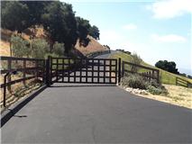 Black gate and fence on a paved driveway leading up a hill with a green and brown landscape.