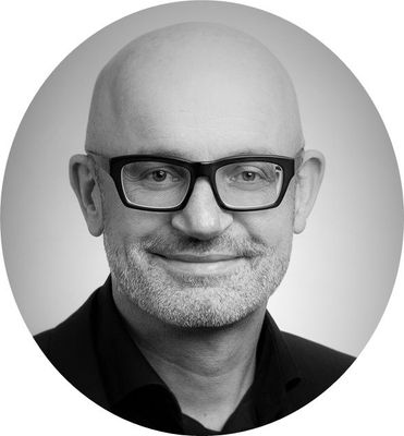 Zwart-wit portretfoto van een kale man met bril en donker shirt, die lichtjes glimlacht.