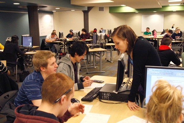 Image of a woman looking over a computer screen in a room of students working at computer stations