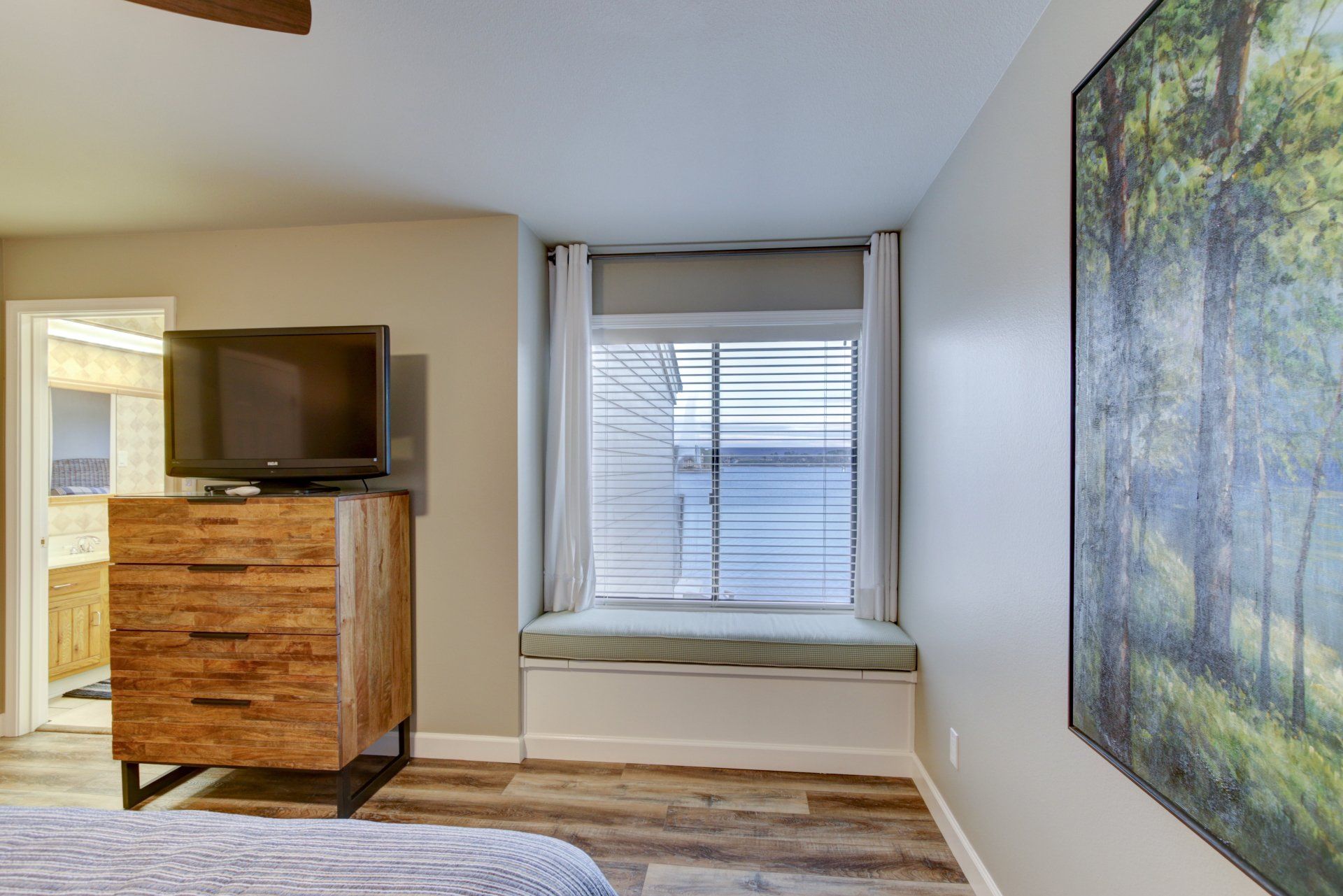 A bedroom with a bed , dresser , television and window seat.