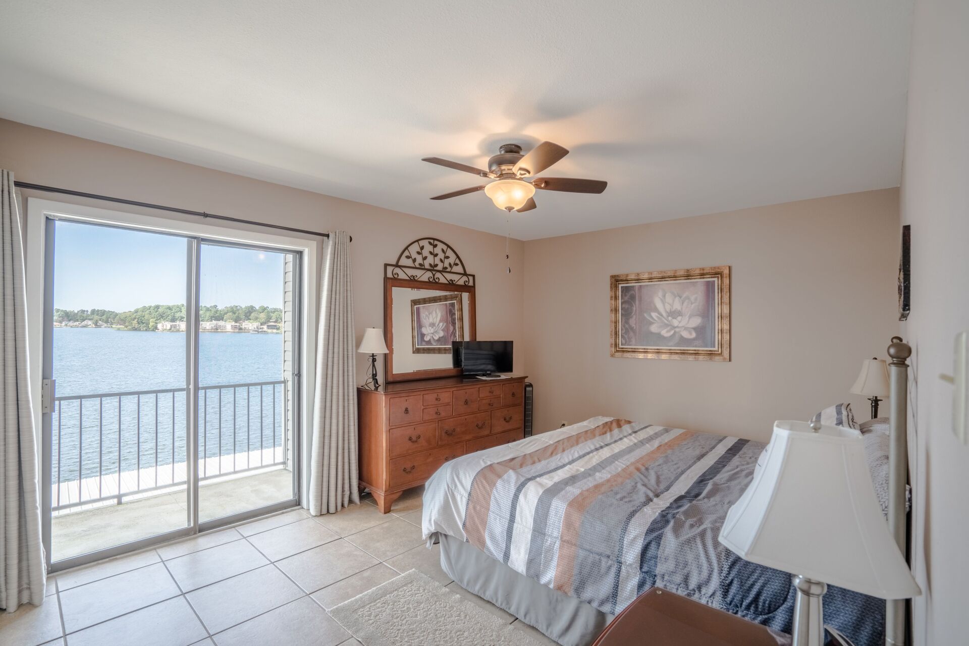A bedroom with a large bed , a ceiling fan , a dresser and a view of the water.