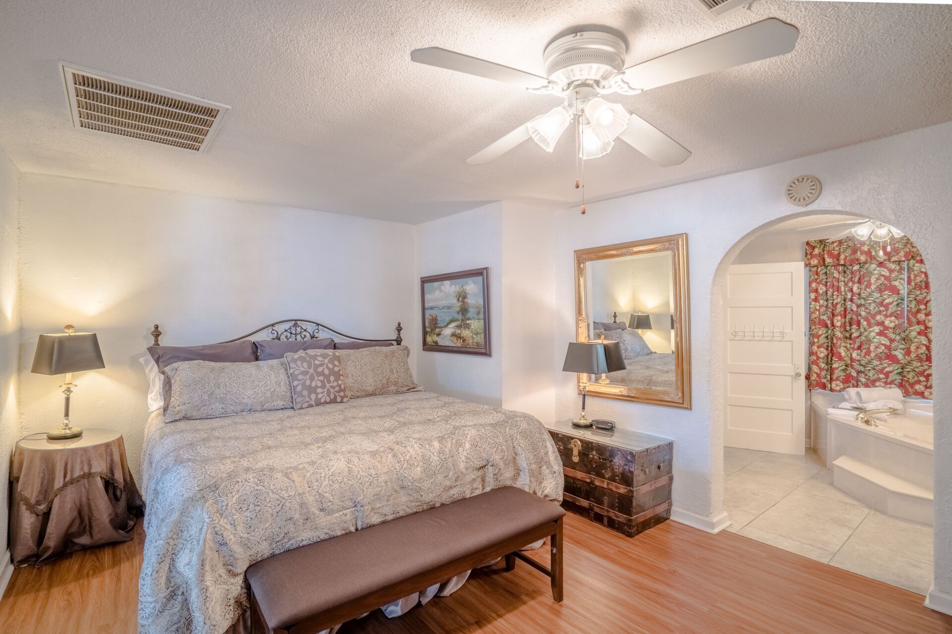 A bedroom with a king size bed , a bench , a mirror and a ceiling fan.