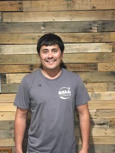 Man with dark hair and grey shirt smiles in front of a wood wall.