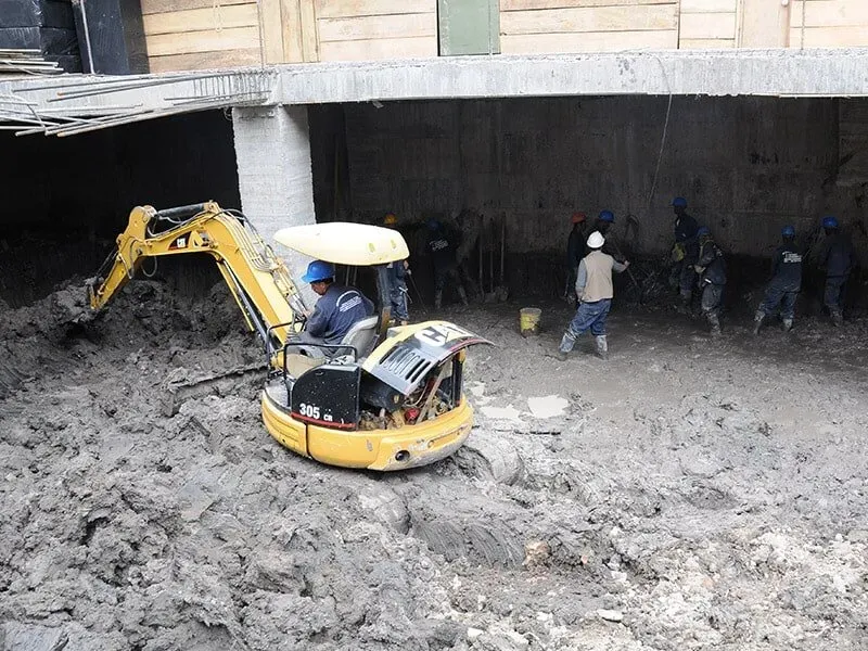 Un hombre conduce una excavadora amarilla en una zona fangosa.