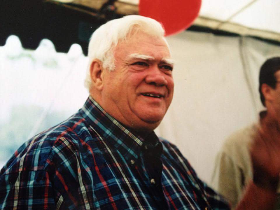 Older white man with white hair and eyebrows smiles; wearing a blue, black, and red plaid shirt.