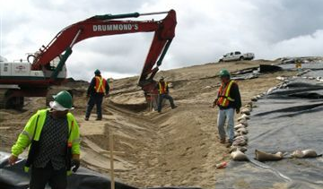 Construction workers on a sandy site with an excavator, laying black tarp, and wearing safety vests and hard hats.