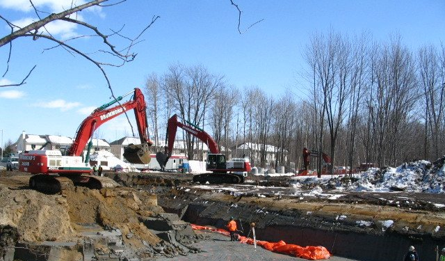 Three excavators digging a trench in a construction site, with snow on the ground and houses in the background.