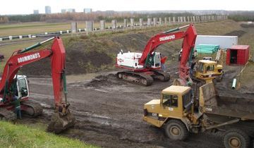 Construction site with red excavators, a yellow dump truck, and a white truck working on a muddy landscape.