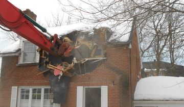 Red excavator demolishing a brick building's roof; snow on the roof and ground.