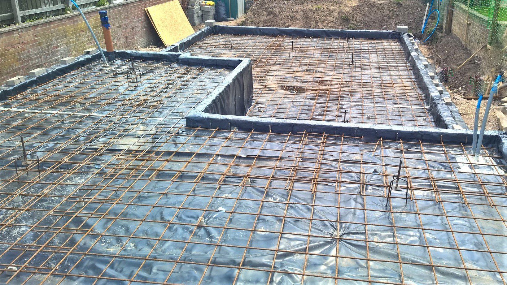 Steel rebar grid laid over a foundation slab under construction, with formwork and gravel around it