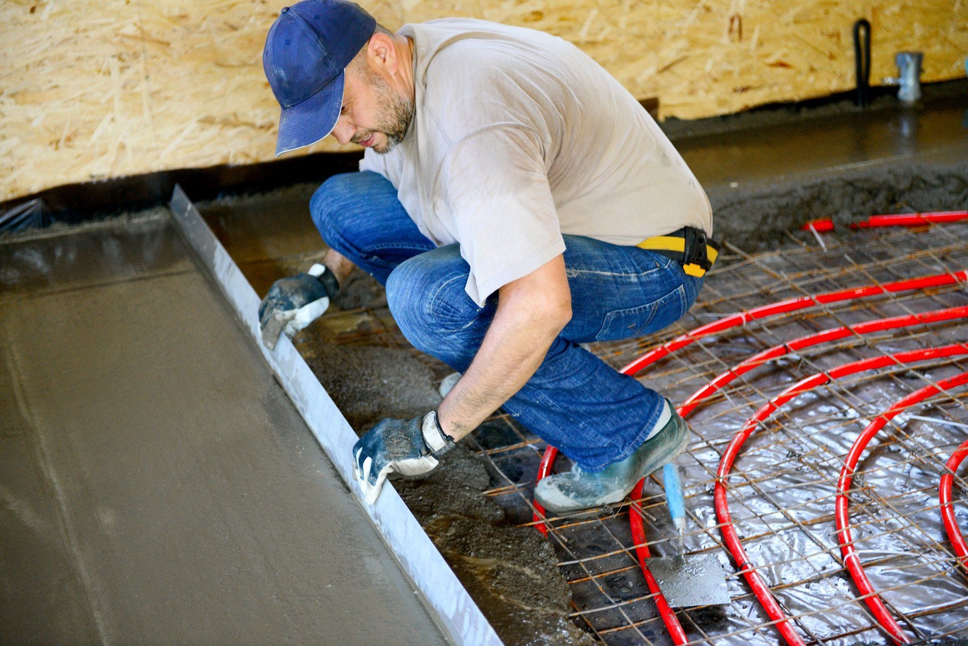 Worker smoothing wet concrete beside red tubing in an indoor construction area
