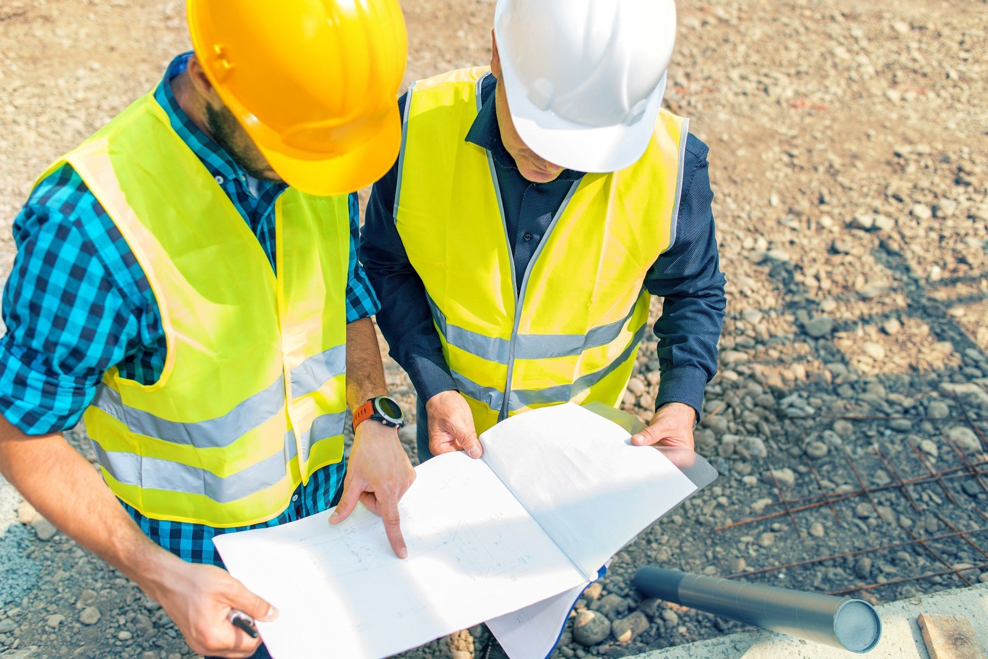 Two construction workers in hard hats and safety vests reviewing blueprints outdoors.