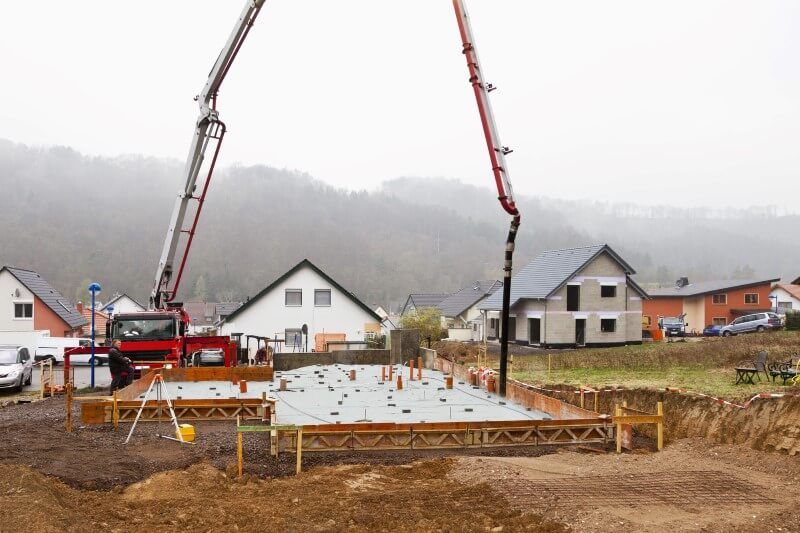Concrete foundation being poured at a house construction site with two red pumps and houses in the background