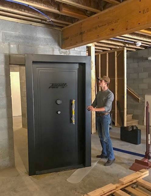 Man standing beside a large black vault door in an unfinished basement
