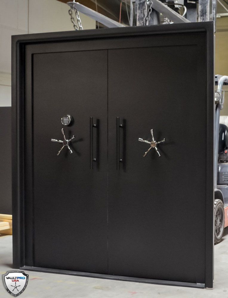 Large black industrial vault door with dual handles and combination wheels in a workshop.