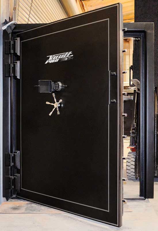 Large black industrial vault door open in a building, showing heavy hinges and locking mechanism.