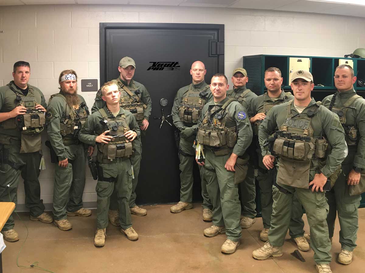 Group of uniformed tactical officers standing indoors in a briefing room, some holding rifles.