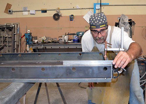 Man in workshop inspecting a metal beam on a workbench, wearing a cap and safety glasses