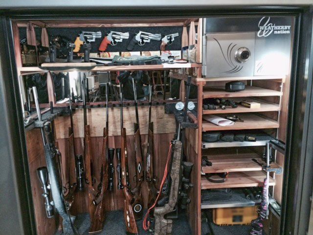 Gun cabinet filled with rifles and handguns, with ammunition and accessories on wooden shelves.