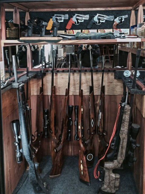 Rifles and shotguns neatly stored in a wooden gun cabinet, with a handgun and gear on the top shelf