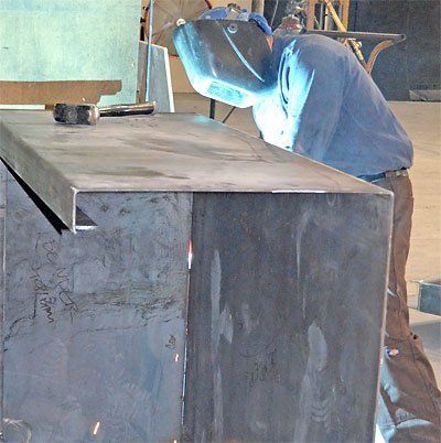 Person welding a large metal sheet in a workshop, wearing a blue shirt and face shield.
