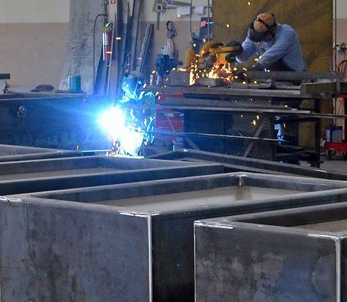 Worker welding metal frames in a workshop, with bright blue sparks and orange grinding sparks nearby