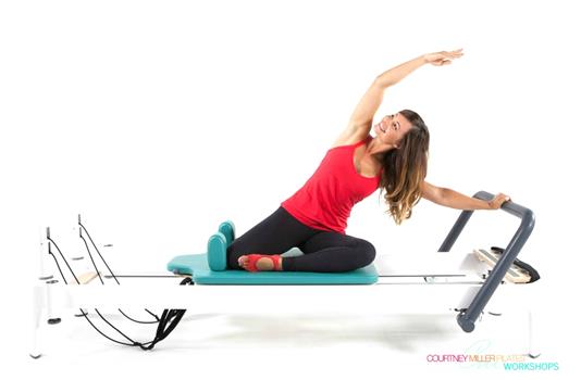 A woman is sitting on a pilates refomer stretching her arms.