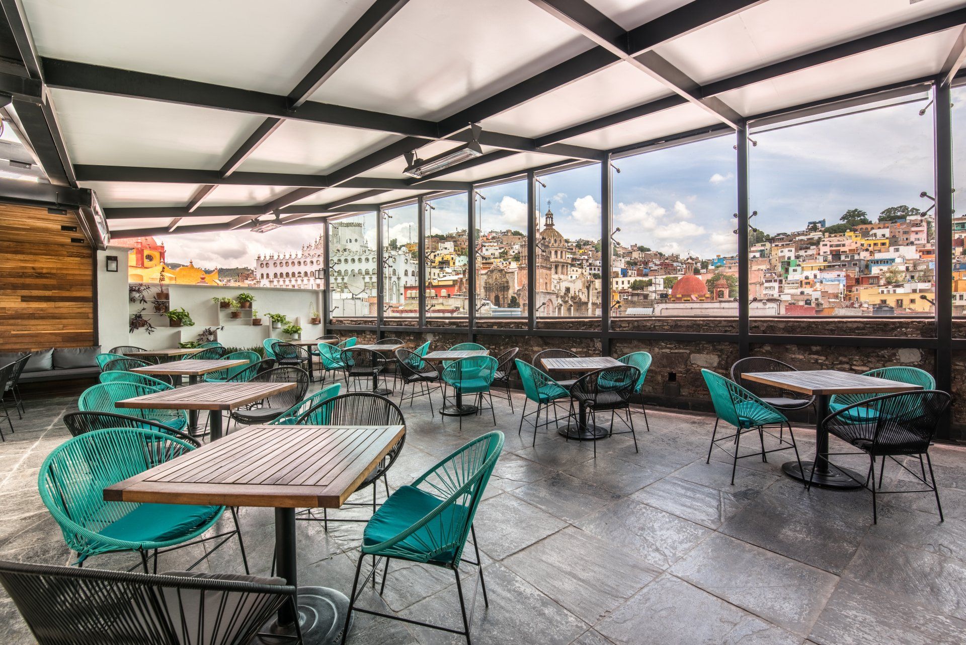 Rooftop café with teal chairs, wooden tables, and city views through large glass windows.