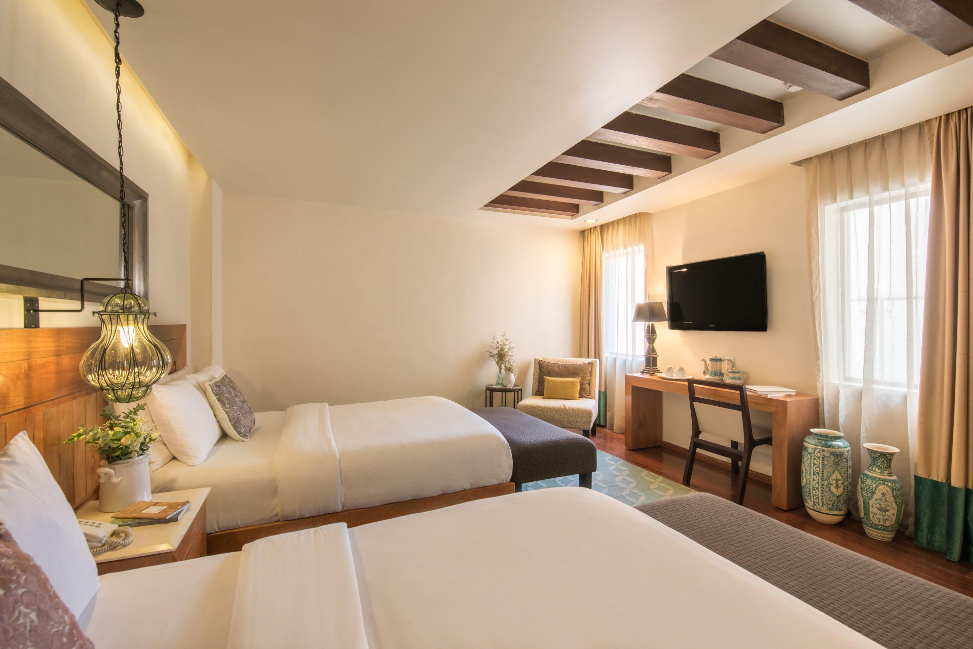 Bright hotel room with two beds, TV, warm lighting, and curtains by a window.