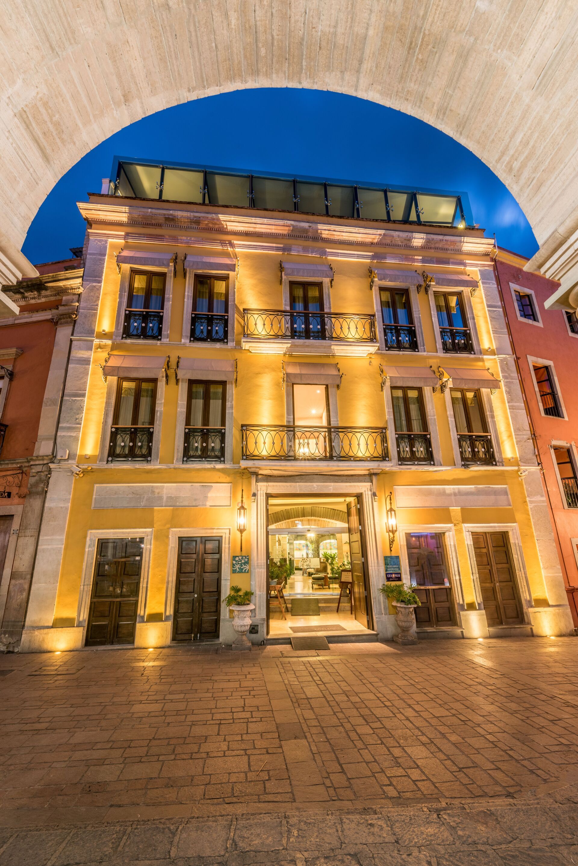 Illuminated historic building façade under an arched stone ceiling at dusk, with a lit entrance courtyard.