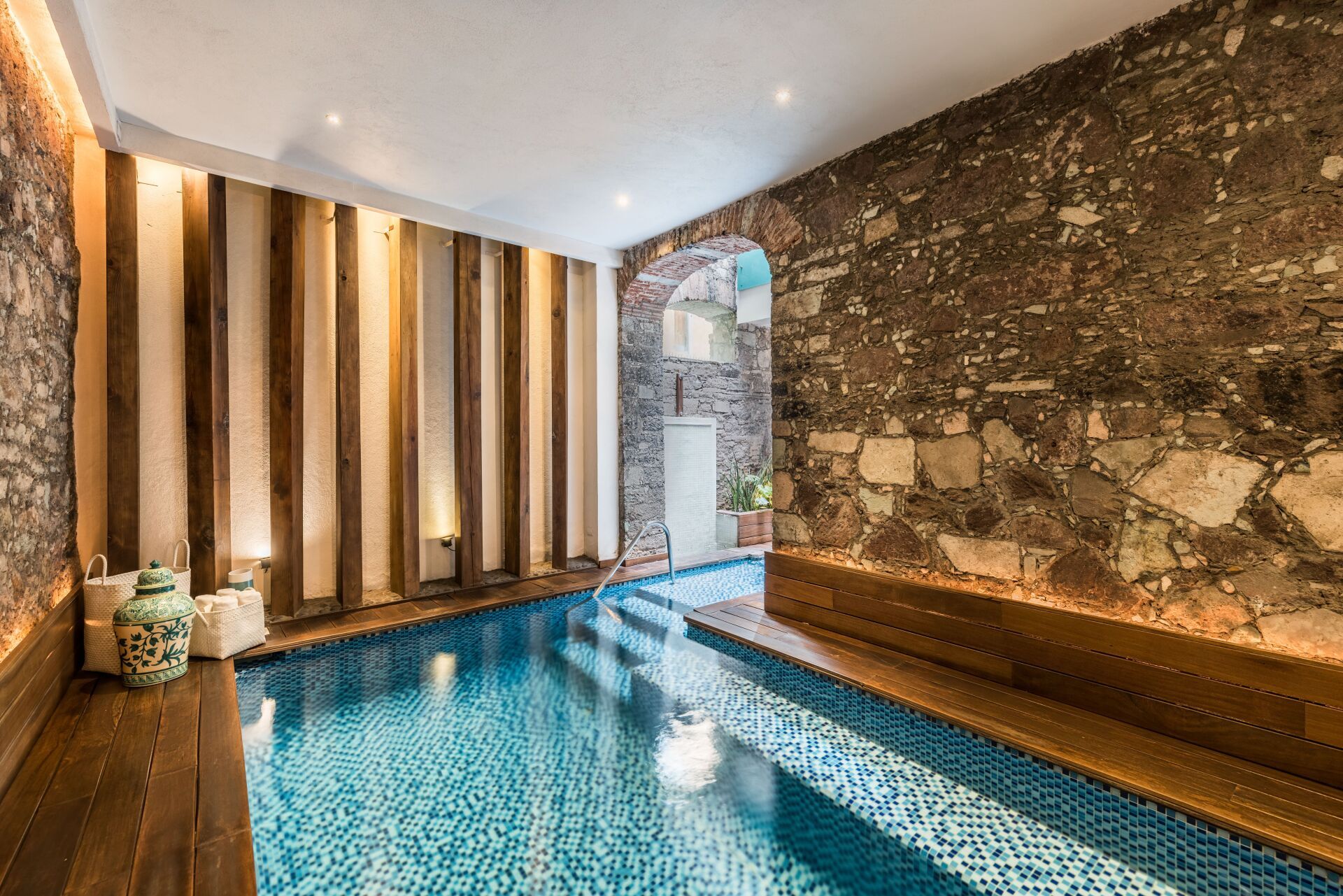 Indoor pool with blue water, stone walls, and striped window shades in a spa-like room