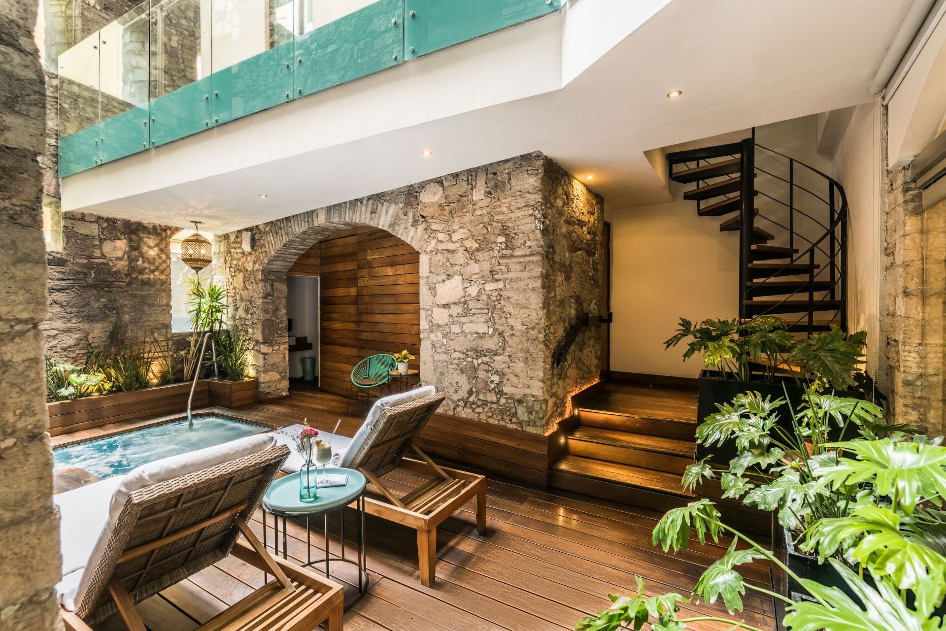 Courtyard lounge with stone walls, pool, wicker chairs, and a spiral staircase under a balcony
