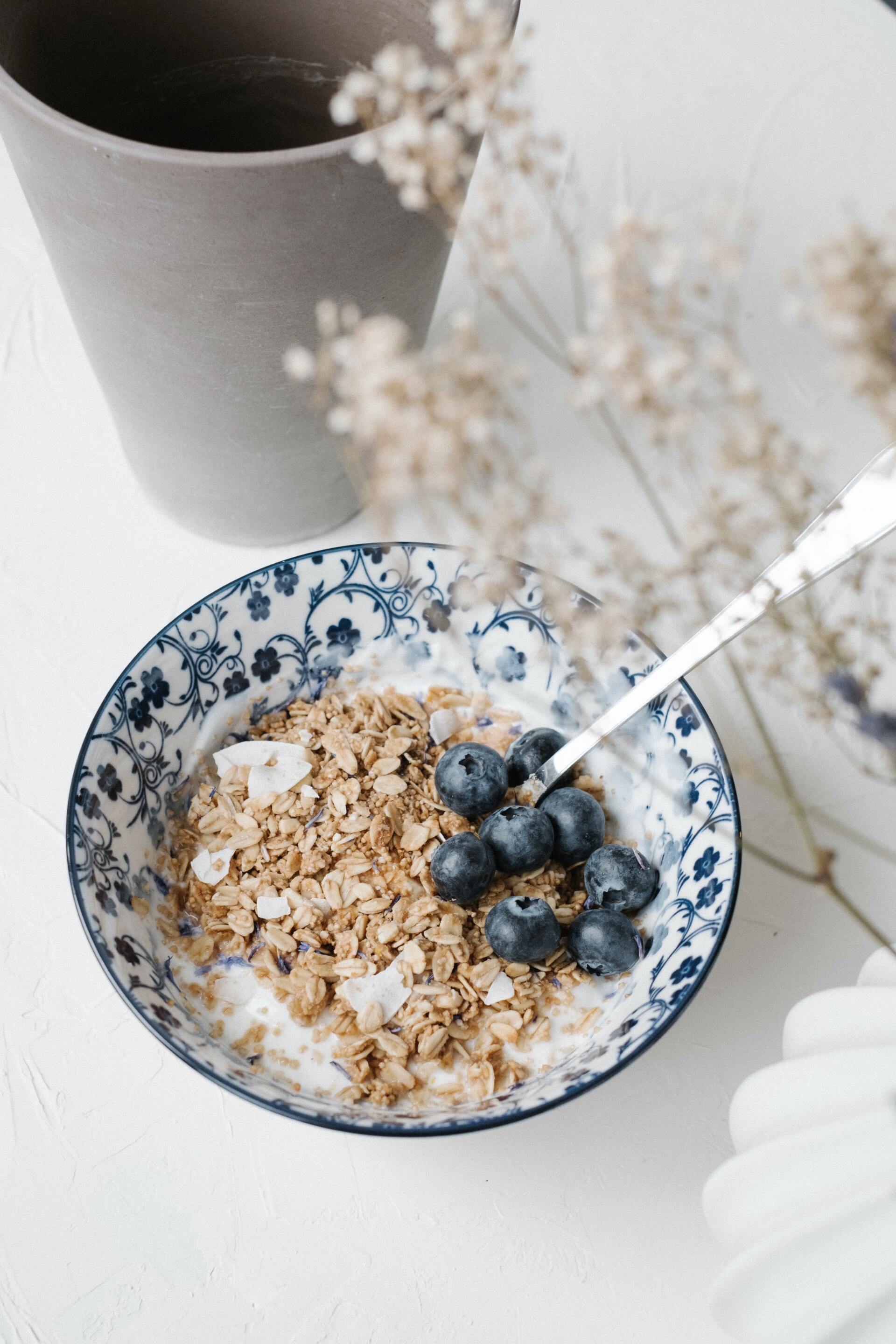 Blue-patterned bowl of cereal with blueberries on a white table, with a cup and dried flowers nearby