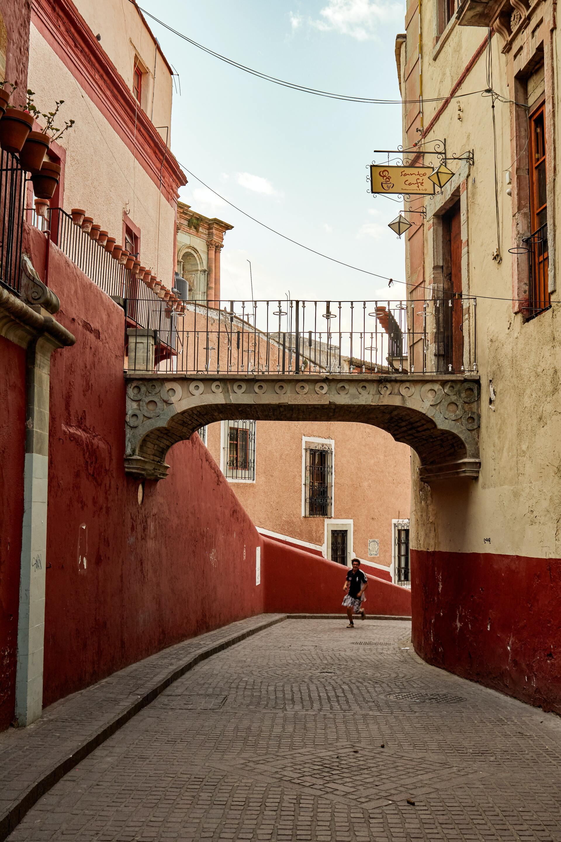 Narrow cobblestone alley with red and cream buildings, overhead bridge, and a person walking ahead