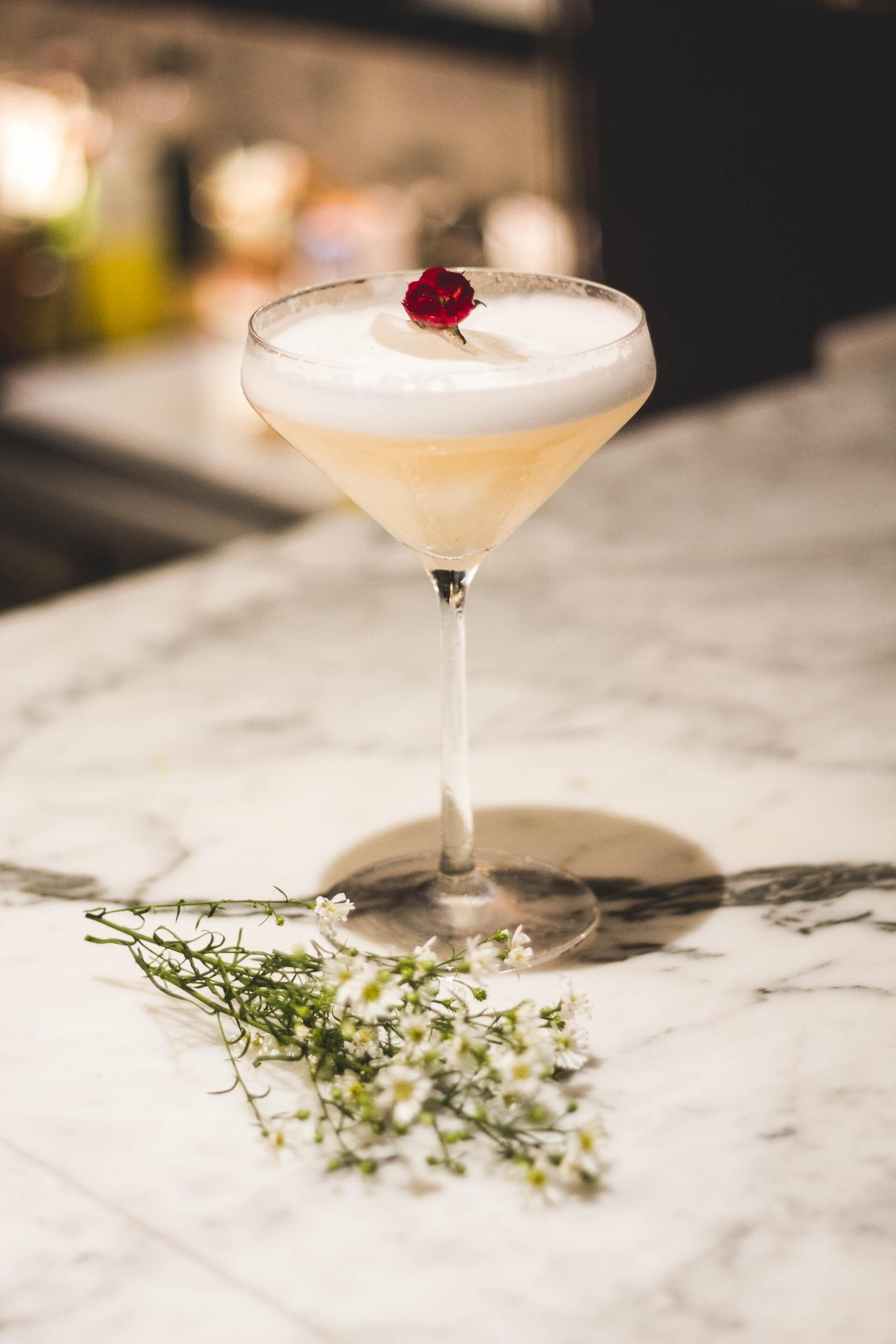 Cocktail in a stemmed glass with a white foam top and red garnish on a marble bar counter