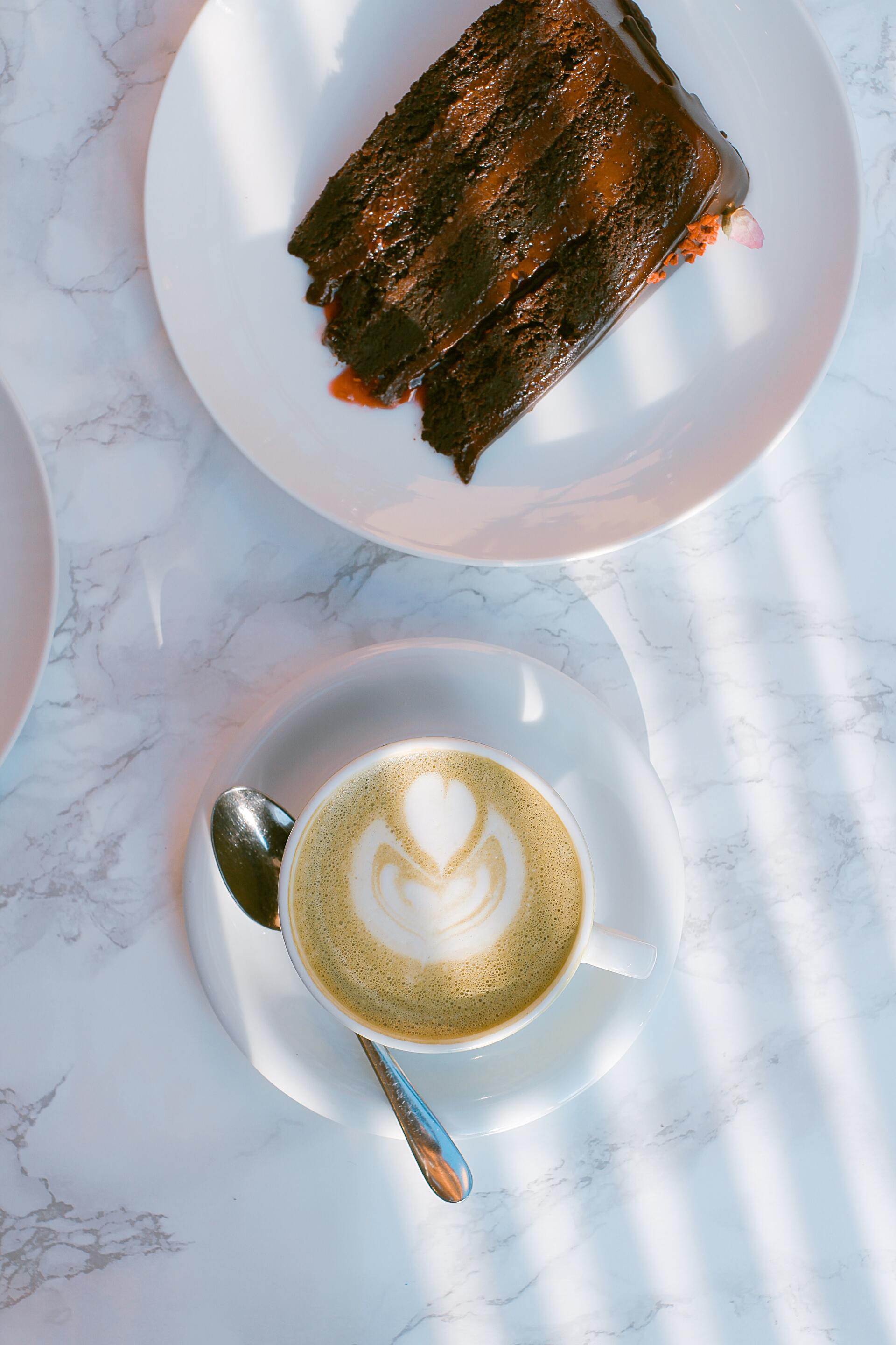Top-down view of a cup of latte, spoon, and a white plate with chocolate-drizzled toast on a marble table