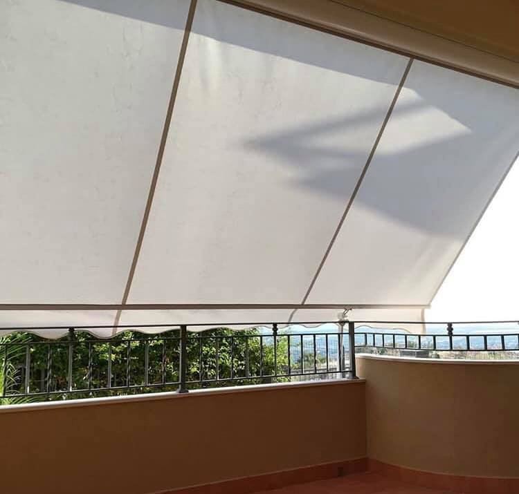Balcone con tenda da sole bianca estesa, ringhiera in metallo e alberi. La luce del sole crea ombre.