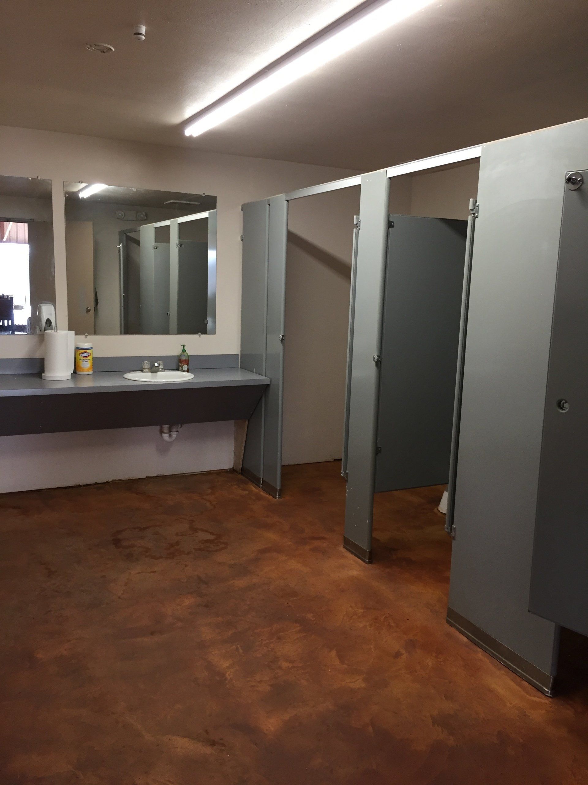 Public restroom with gray stalls, brown floor, and a long sink counter.