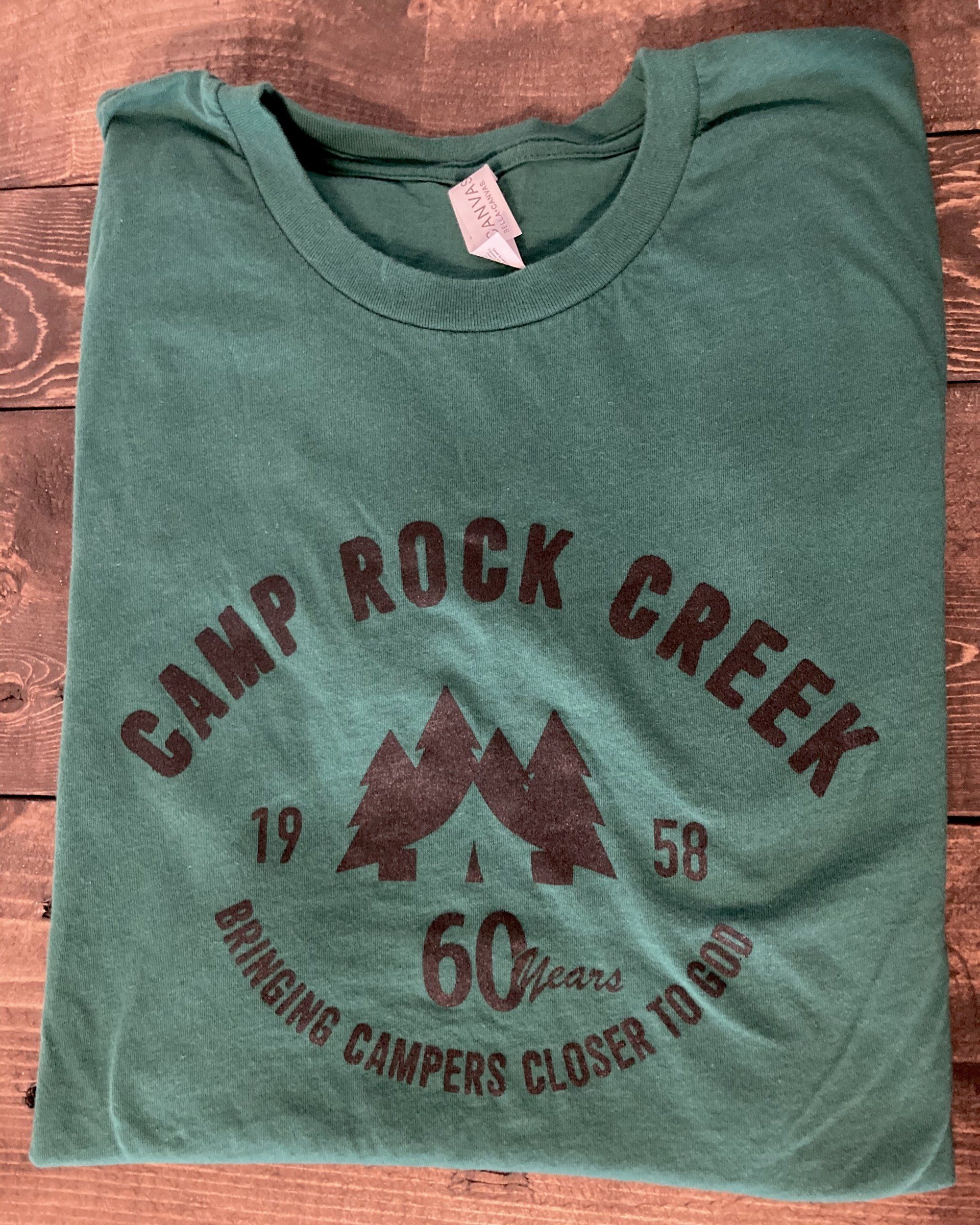 Green Camp Rock Creek t-shirt with black text and logo, folded on a wooden surface.