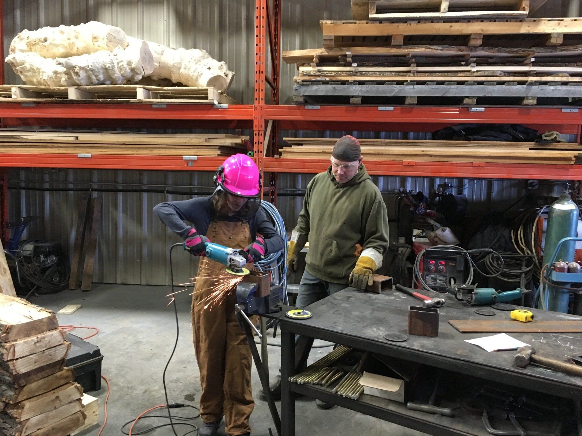 Two workers welding metal in an industrial workshop, one wearing a pink hard hat near stacked materials.