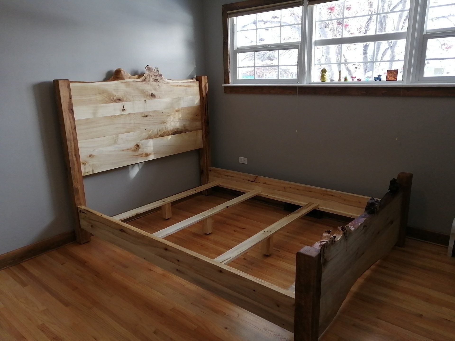 Partially assembled wooden bed frame in a sparsely furnished room with hardwood floor and window.