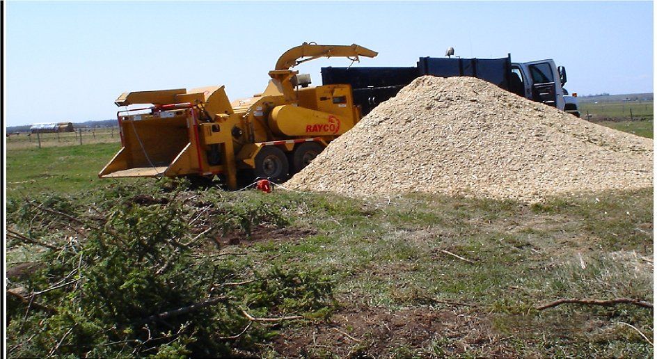 Yellow wood chipper beside a large pile of sawdust on a rural worksite, with a truck in the background