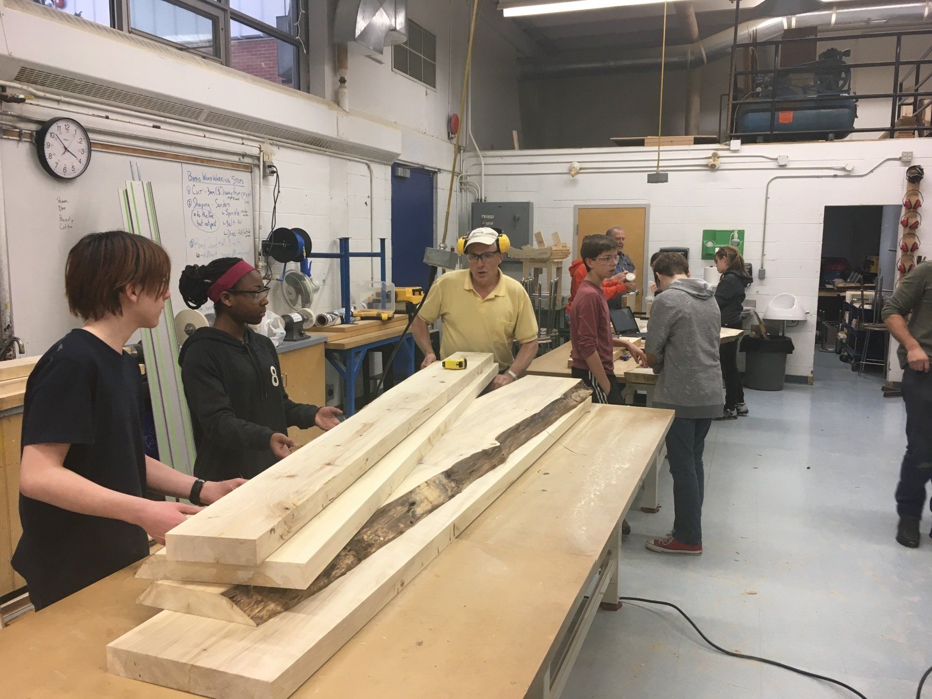 People gathered around long wooden planks in a workshop, discussing or handling materials.
