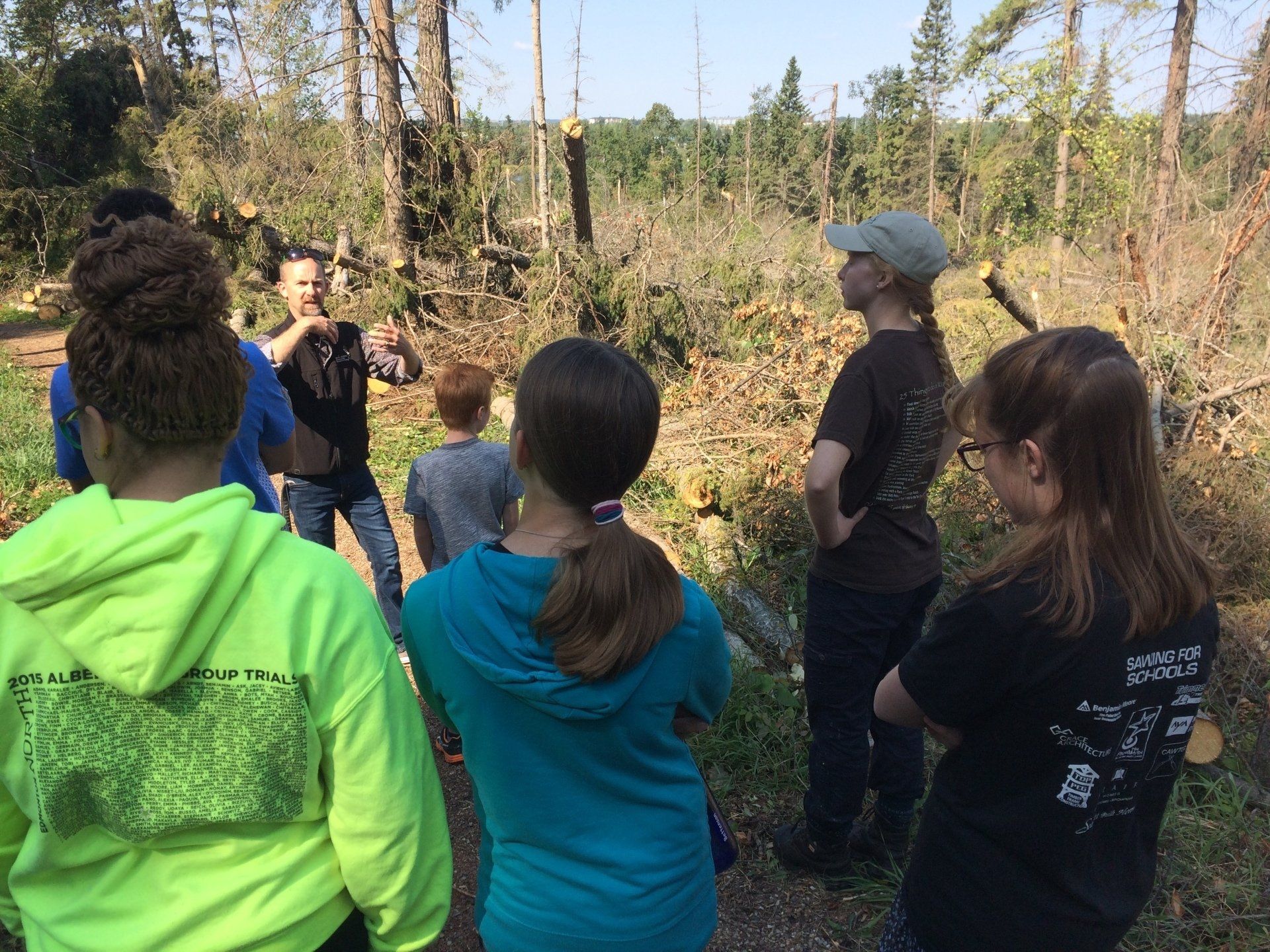 Group of people listening to a speaker outdoors in a wooded area, with one person gesturing near the center.