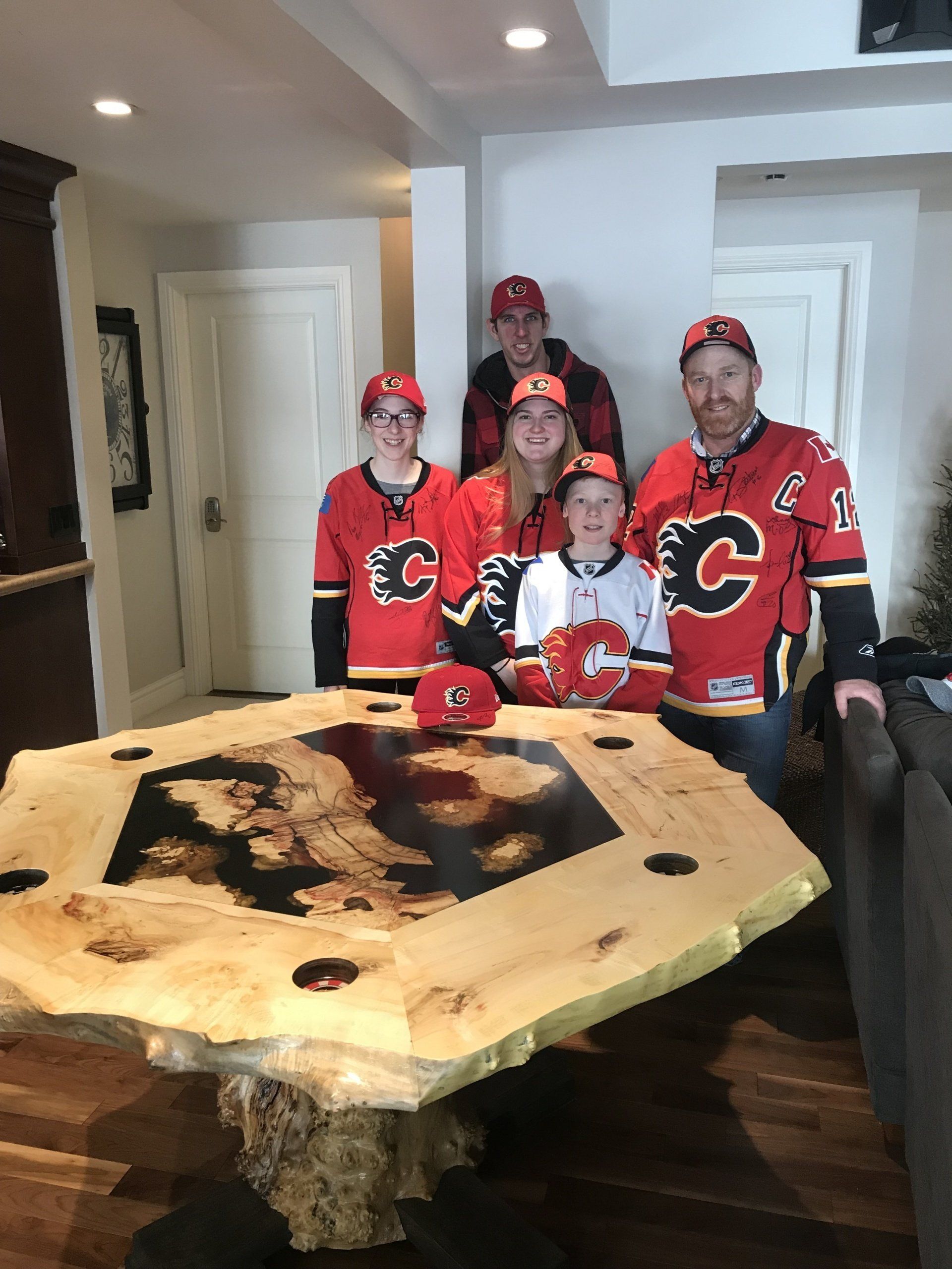 Four people in red hockey jerseys stand behind a large live-edge wooden table in a home interior.