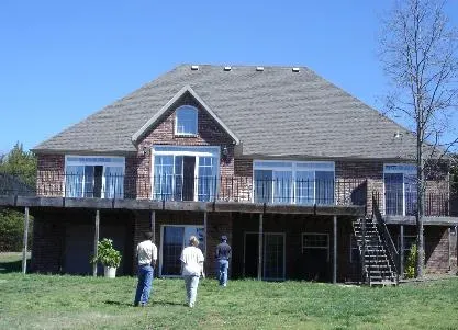 Two people walking toward a large brick house with a raised deck and stairs in a grassy yard