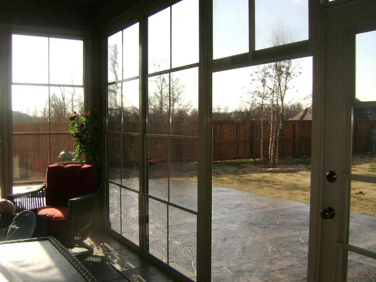 Sunlit enclosed porch with glass doors and windows overlooking a backyard and wooden fence.