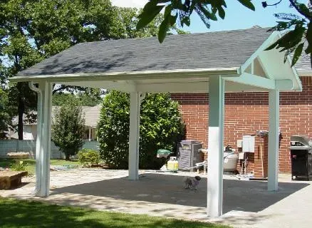 Covered backyard patio with white posts, gray roof, and brick wall beside a house.