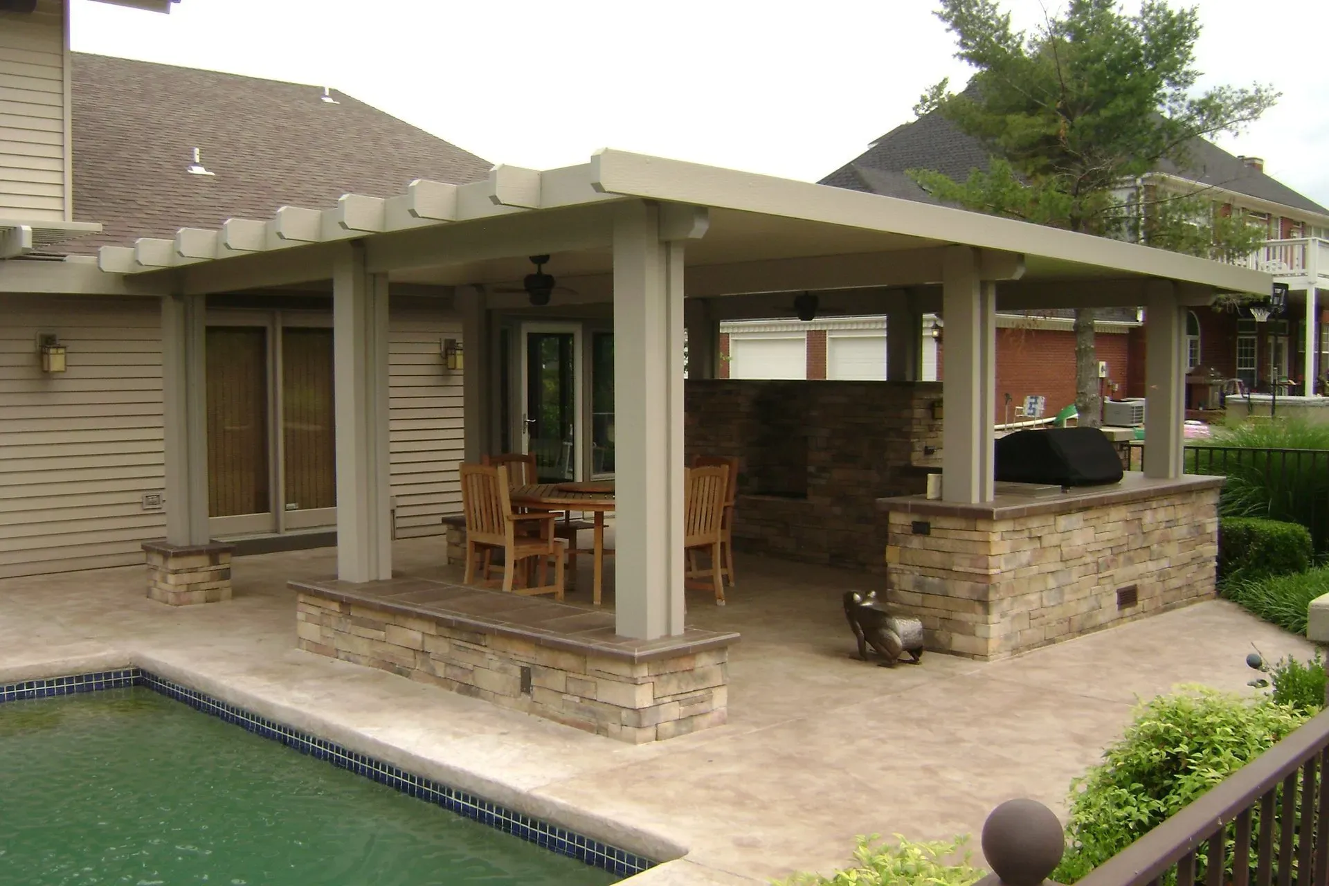 Covered backyard patio with stone barbecue and outdoor dining area beside a swimming pool