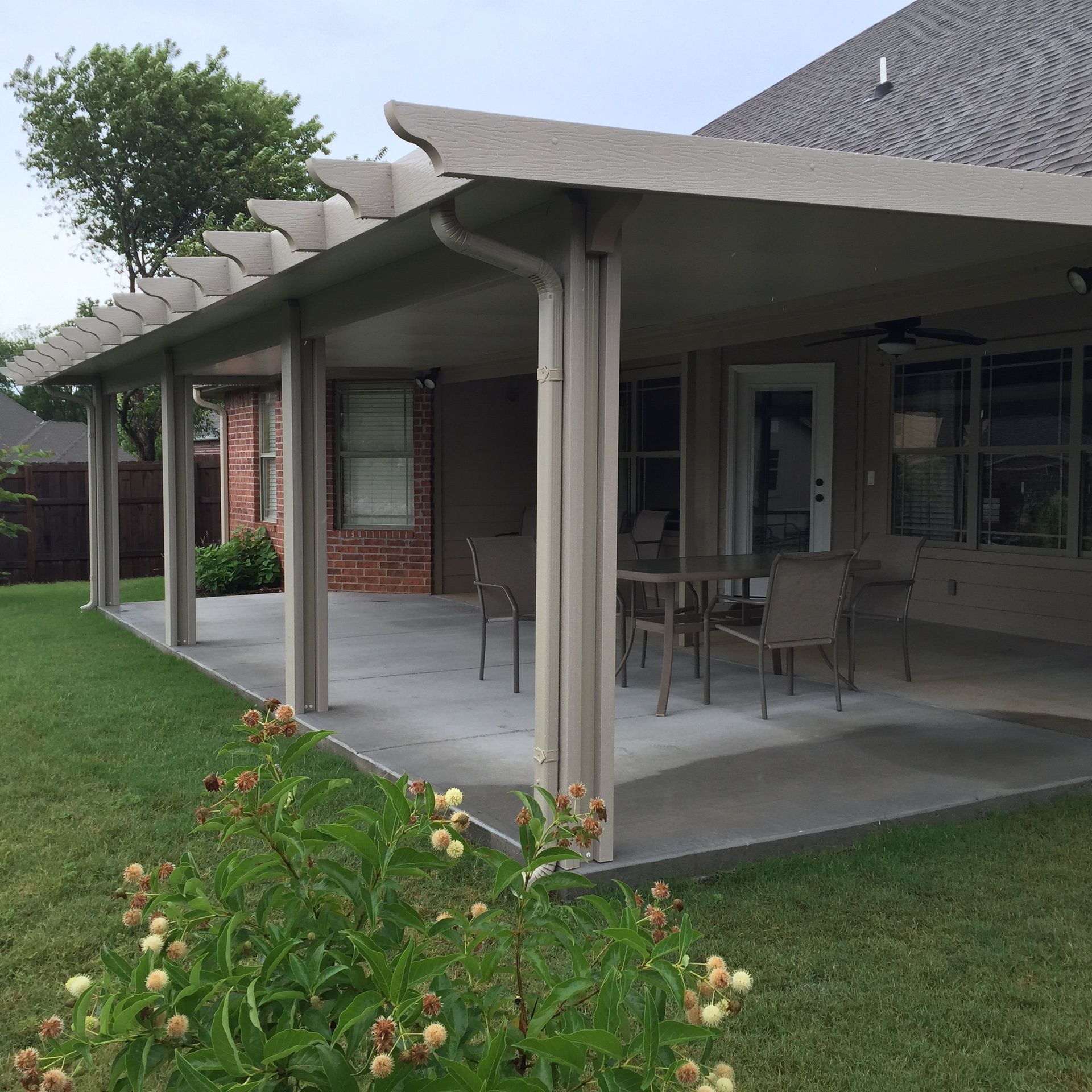Covered patio with outdoor dining table and chairs beside a brick house, overlooking a green lawn